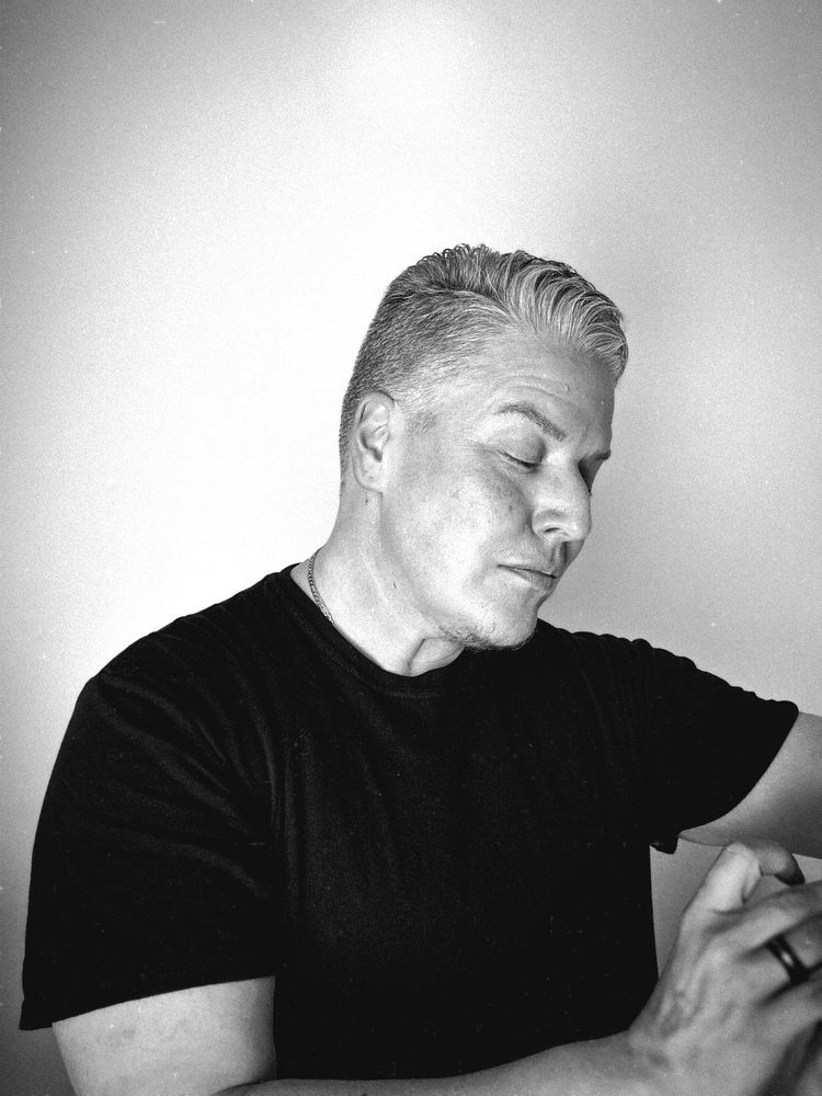 Rowan stands in a black t-shirt looking down at his watch. The photo is in black and white and has a moodiness to it. 