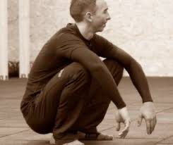 A white appearing man in black clothes sits in a relaxed position, feet flat on the ground, knees bent all the way, butt a few inches off the ground, arms relaxed over his knees. Is this sitting in a squat?