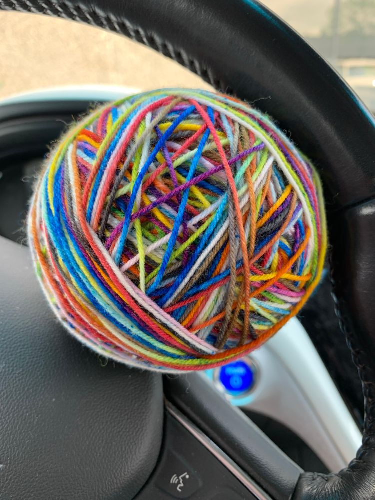 A ball of yarn in the colors of the progress pride flag is resting in a steering wheel of a parked car.