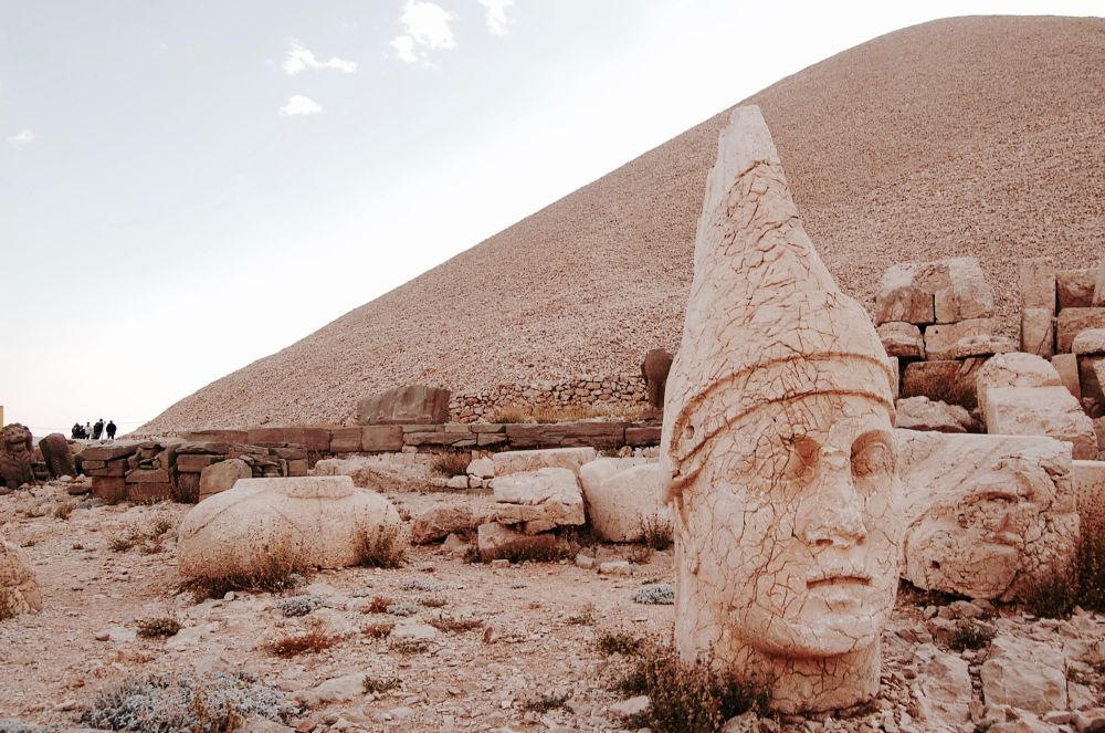 Huge statue head of Apollo-Mithra-Helios-Hermes located on the West terrace of Mt. Nemrut in Turkey. The head shot is on the right side of the photo and behind it is mountain of what is known as a royal tomb and big slabs of stone. On the left side of the photo is a tiny shot of people or tourists. It's a UNESCO World Heritage Site and one of my most unforgettable travels.