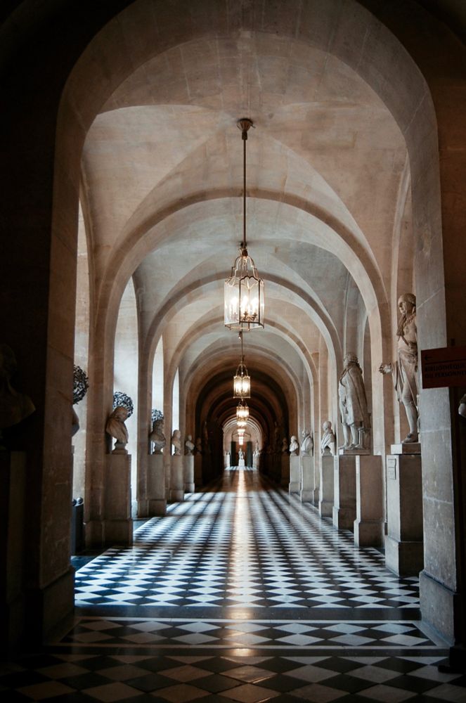 An arched hallway lined with marble statues. Black and white argyle tiled floors. Dimmed hanging lights in the middle of each archway. Long shot showing the length of hallway highlighting the symmetry of the light reflection on the floors. Taken at the Versailles palace.