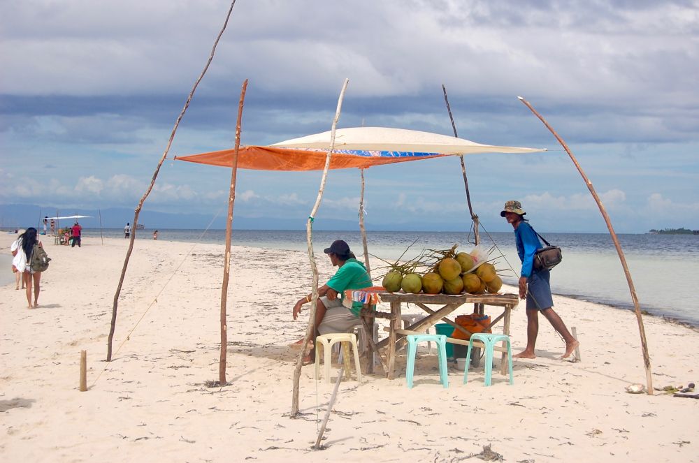 A tropical scene of a coconut vendor, a man wearing a green tee, sitting under a makeshift tent on a sandbar looking at tourists walking by, with a bunch of coconuts on a table and some monoblock chairs. There is another man in blue long-sleeves shirt carrying a black bag and wearing a hat, seemingly looking at the camera (me!), he is barefoot. The blue, cloudy skies is a bit overcast and evokes a mood of impending rains. This shot was taken in Bohol island.