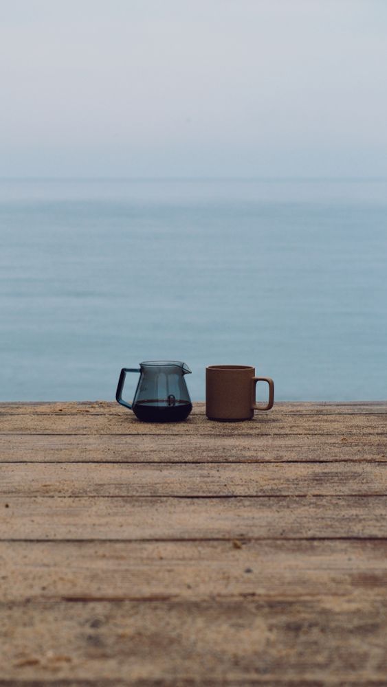 The photo shows a glass carafe with coffee and a beige porcelain mug at the end of a walkway overlooking the ocean on a foggy morning on #capecod