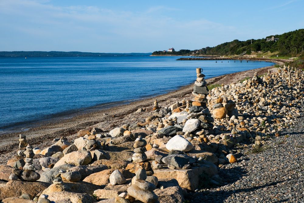 The photo shows an area where people stack rocks on top of each other to form cairns, beside the ocean in Falmouth, MA on Cape Cod. On the tallest pile of rocks is a mug, taking the place of the top rock. The mug is beige porcelain and matches the hues of the rocks all around. The ocean is in the background, with Martha's Vineyard clearly visible in the distance on the left and the Nobska Lighthouse Keeper's  House visible in the distance in the center right. 