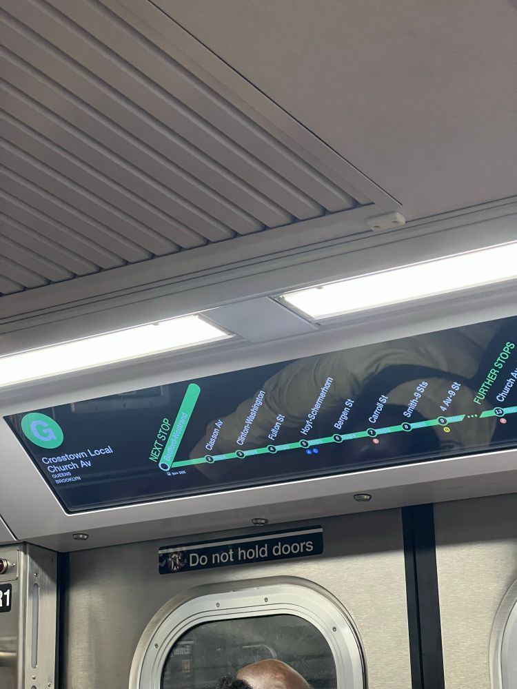A screen in a subway car showing the route of the train. The first stop is highlighted with a light green background but has white text that is barely readable against the green background.