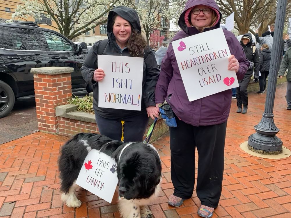 Two women holding protest signs with a dog wearing a sign. 