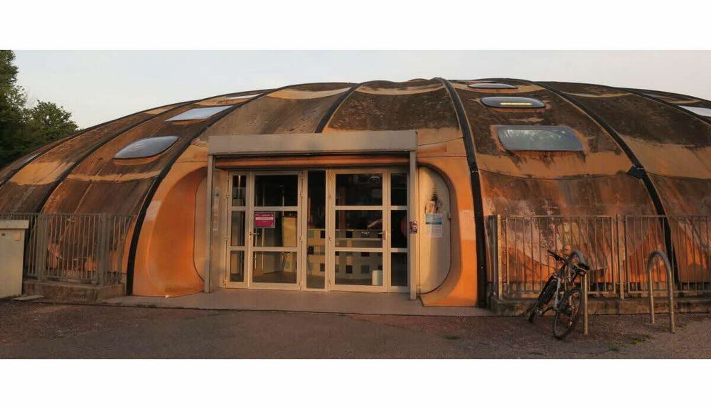 Encore une piscine tournesol bien crasseuse, mais en version orange. On aperçoit l'accueil à travers les portes vitrées de l'entrée. Il y a un vélo attaché devant et quelques affiches près des portes, laissant supposer qu'elle est encore en activité malgré son état.