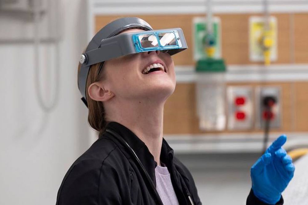 Press photo of Taylor Dearden as Mel King on The Pitt, she's wearing a visor with magnifying glasses and looking up at the ceiling and smiling.