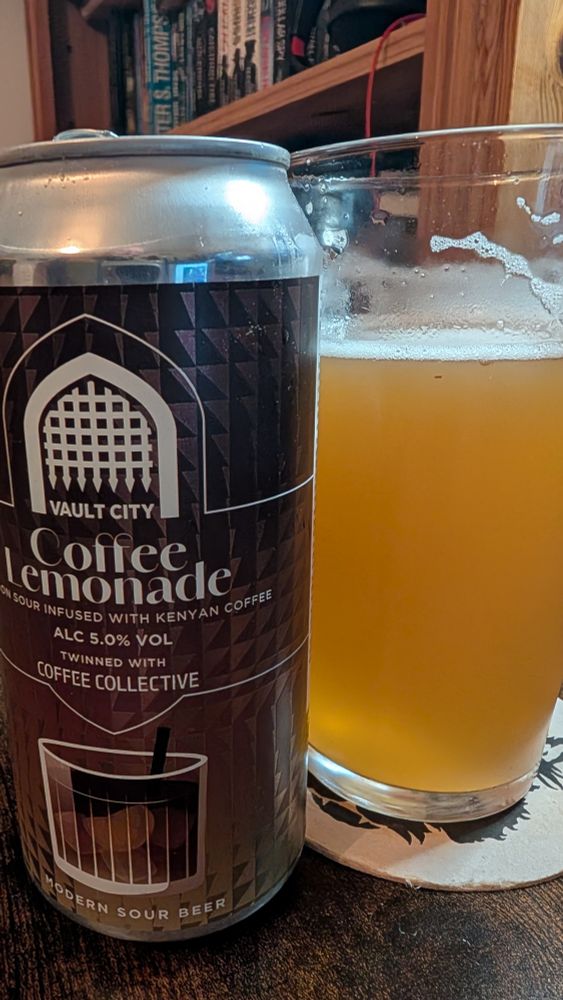 Photograph of a can of Vault City Coffee Lemonade beer next to a glass of the same hazy, light orange beer with a white foam head. The can label is dark brown with a geometric pattern and a white arched window graphic. Text on the label reads: "VAULT CITY Coffee Lemonade LEMON SOUR INFUSED WITH KENYAN COFFEE ALC 5.0% VOL TWINNED WITH COFFEE COLLECTIVE MODERN SOUR BEER". An illustration of a glass with ice and a straw is at the bottom of the label. The beer glass rests on a light-colored coaster with a dark, jagged design. A wooden table and a bookshelf with books are in the background.