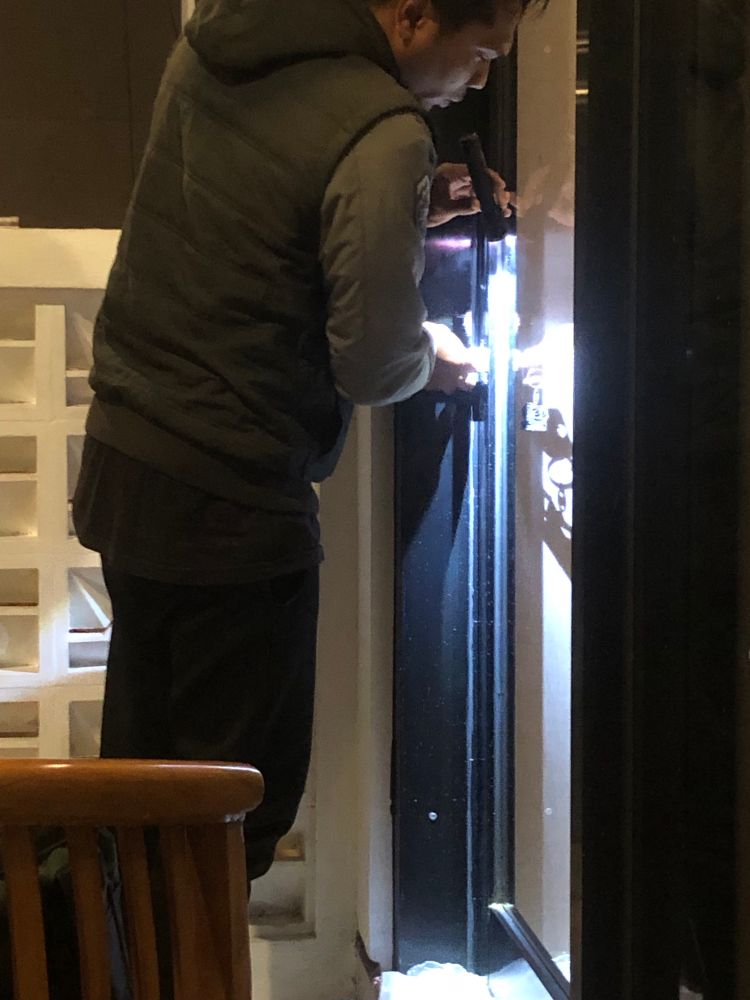 A man stands before a glass door with a flash light. A wooden chair is visible.