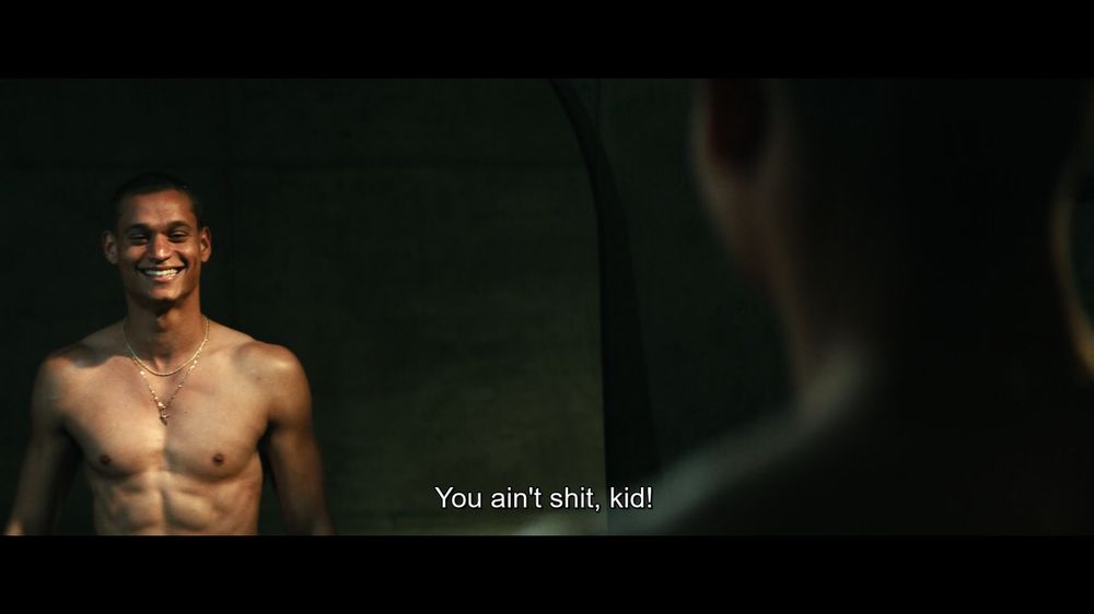 A man grins at his shirtless reflections. A voice says "You ain't shit, kid!"
