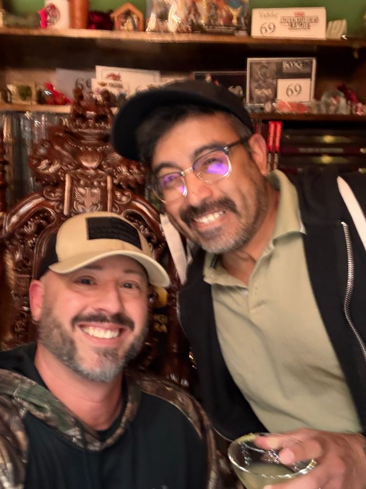 Juan and I at the Gamehole, the home of Gamehole Con. I sit in the DM throne (that’s a real thing) while Juan stands nearby with an adult beverage. A complete D&D library is shelved impressively behind us.