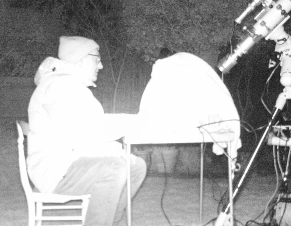 Infrared image of man sat in front of a laptop and telescope, dressed for the chill.