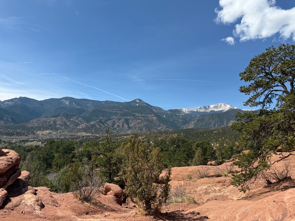 Garden of the dogs in Colorado Springs. Some junipers and other trees are growing in the foreground with a background on the Rocky Mountain range as seen from the east. One mountain has a small snow covering at its peak.