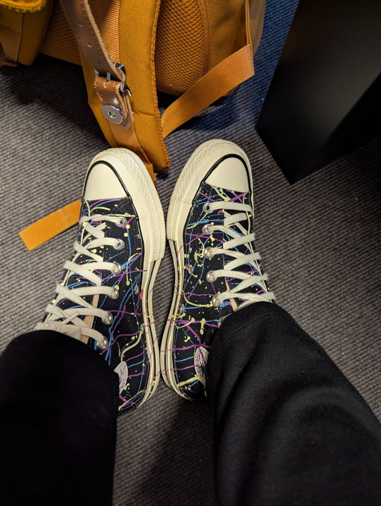 Paintsplatter allstars onder een zwarte broek. De vloer is grijs en er staat een oranje rugzak naast de schoenen.