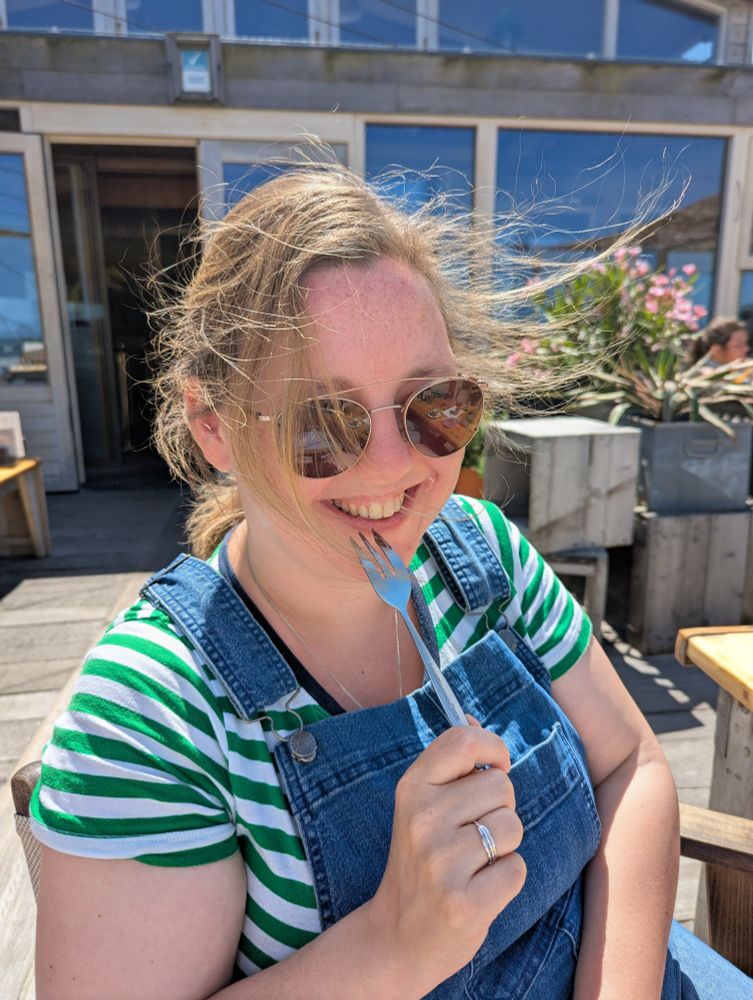 Foto van Anke in groenwit gestreept shirt en denim tuinbroek. Anke heeft haar haar in een staart maar er waaien plukken uit. Ze zit op een terras en veel is van hout. Ze houdt een boekje bij haar mond en lacht, zonnebril op. 