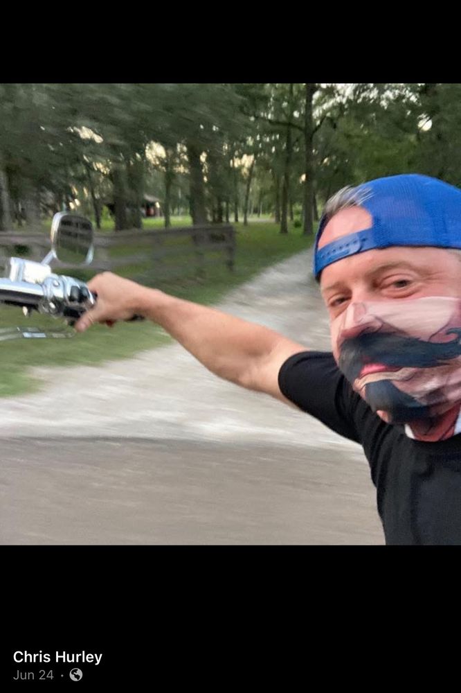 Chris Hurley, a white man, wearing a mask that has a mustache and goatee design while riding a motorcycle.