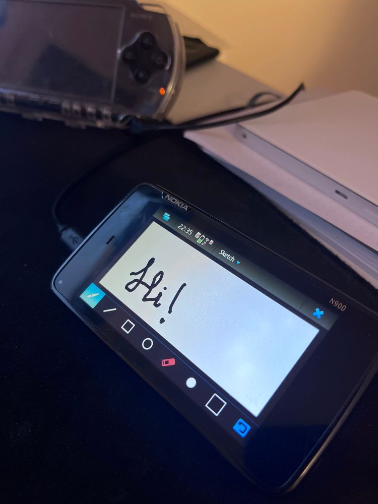 Nokia n900 with the Sketch app open. There is a cursive “Hi!” written. In the background there are other electronics. 