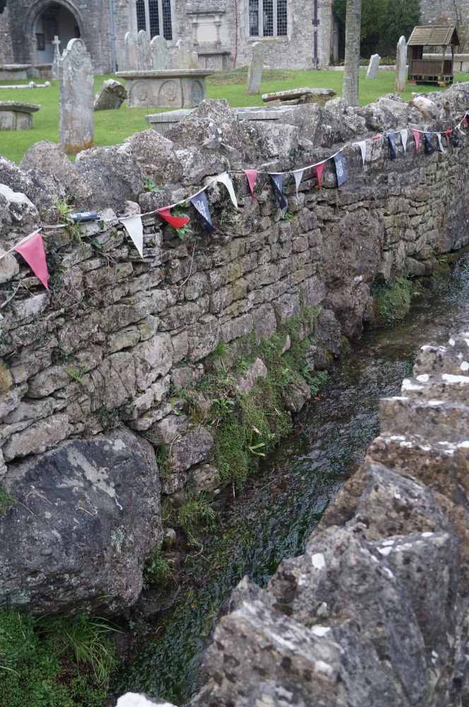 A stone wall, mostly built with small bricks but some large sarsen boulder can be seen lower down. There is a stream in front of the wall and a graveyard visible behind