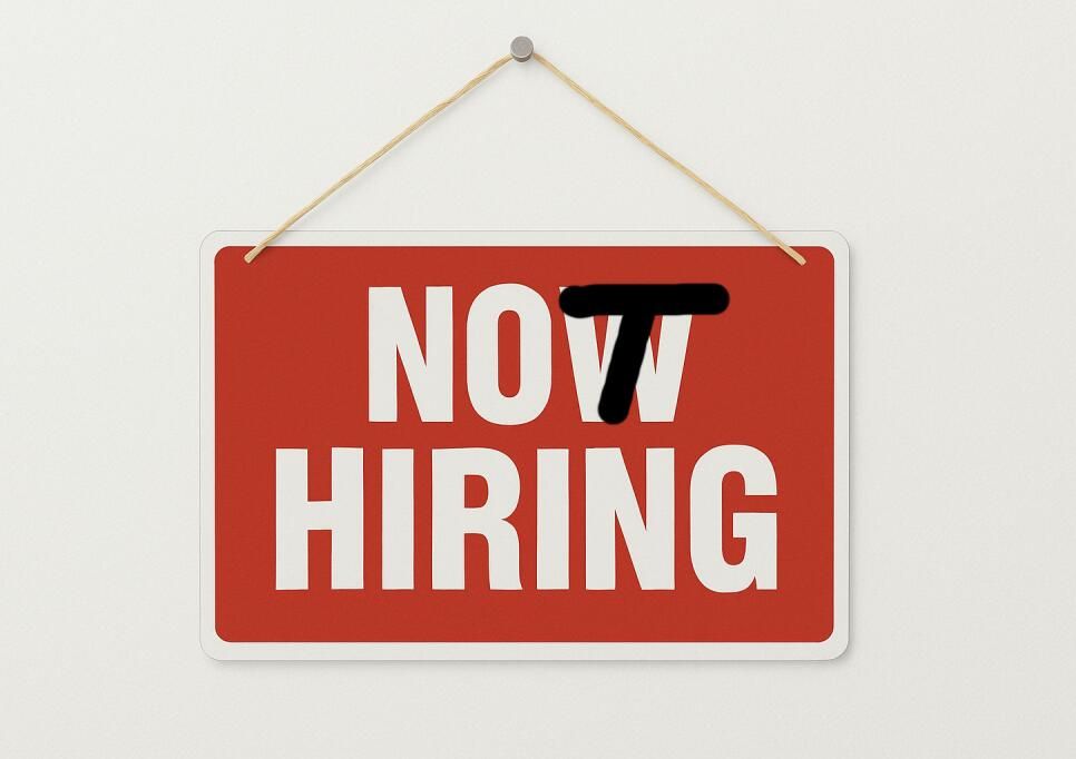 A high-contrast image featuring a red rectangular sign hanging on a plain white wall by a thin piece of twine. The sign has the large white text "NOW HIRING". The word "NOW" has a thick, black horizontal line crudely drawn over the letter 'N' and part of the 'O', changing the meaning to imply "NOT HIRING". The style is simple and graphic, with bright colors and a clear, ironic context about a change in employment status.
