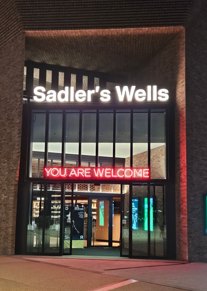 The entrance of Sadler's Wells East theatre. A glass frontage surrounded by brick, with white lights spelling out 'Sadler's Wells' and underneath, red, neon capitals stating 'you are welcome.'