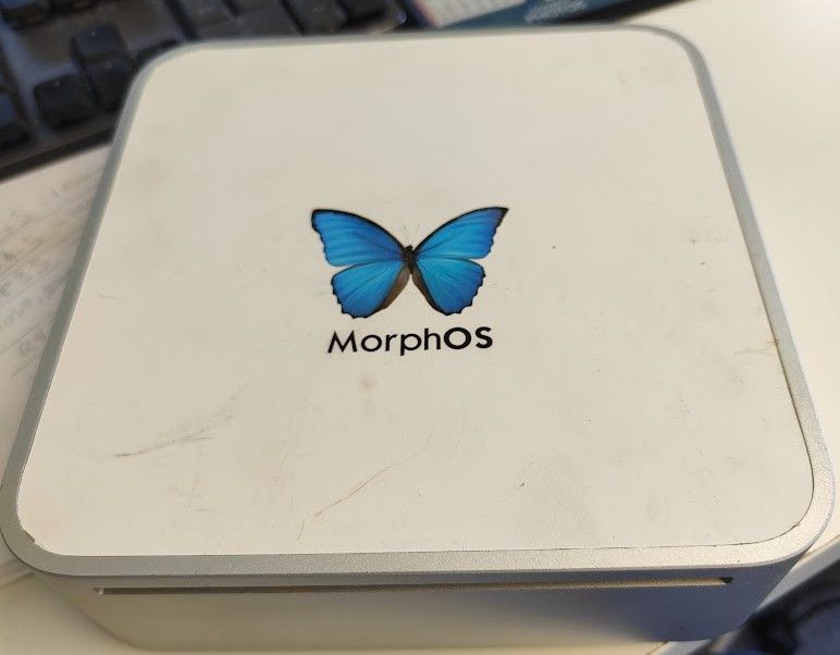 A Mac Mini G3 computer with the Apple logo on the top replaced with a blue Morphos butterfly and text label on a white background.