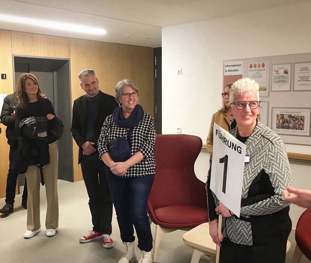 Vier Personen stehen in einem Raum mit zwei Sesseln und schauen eine Person an.