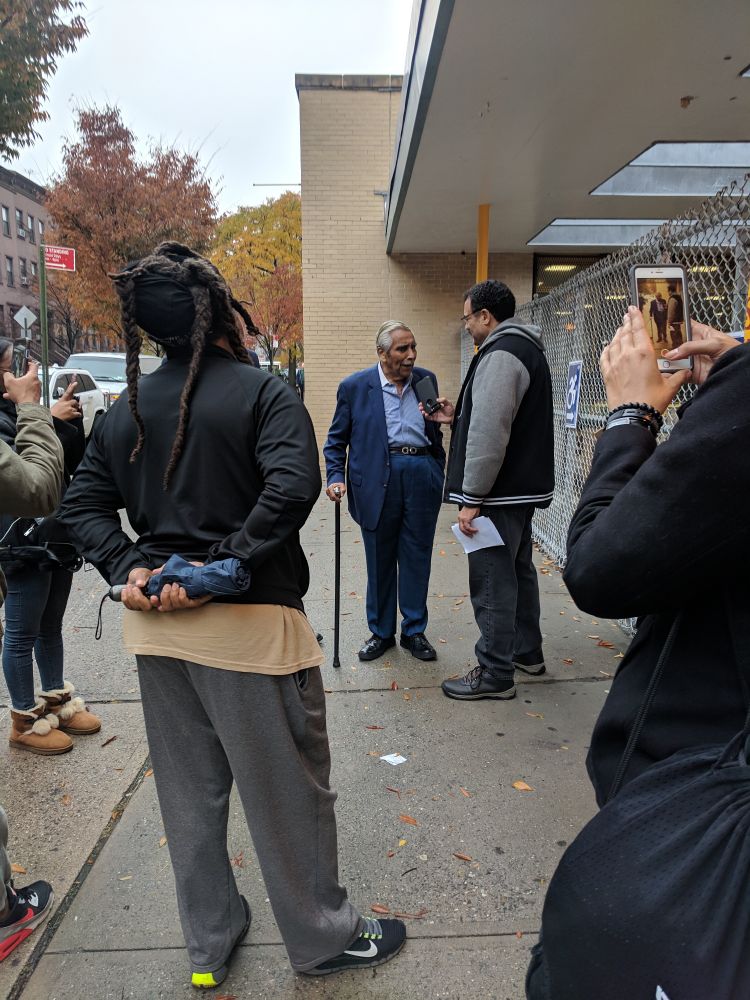 A photo of 88-year old Charles Rangel standing with a cane outside a school, surrounded by a small group of people and being interviewed by a taller man. 