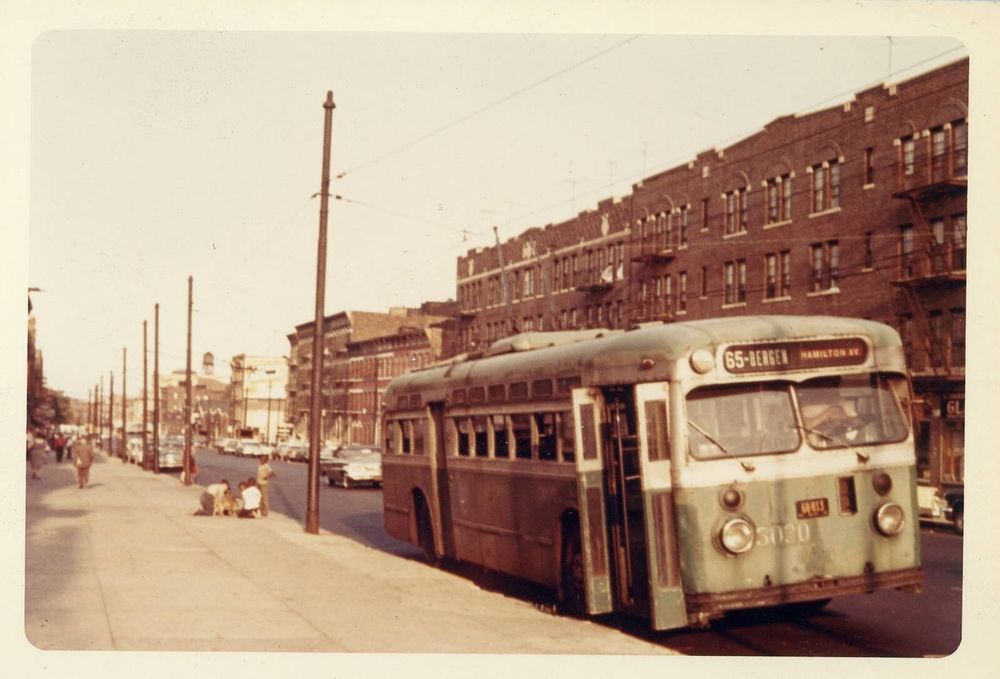 An electric trolley bus in Brooklyn in 1960, with overhead power lines in the foreground and apartment buildings in the back.