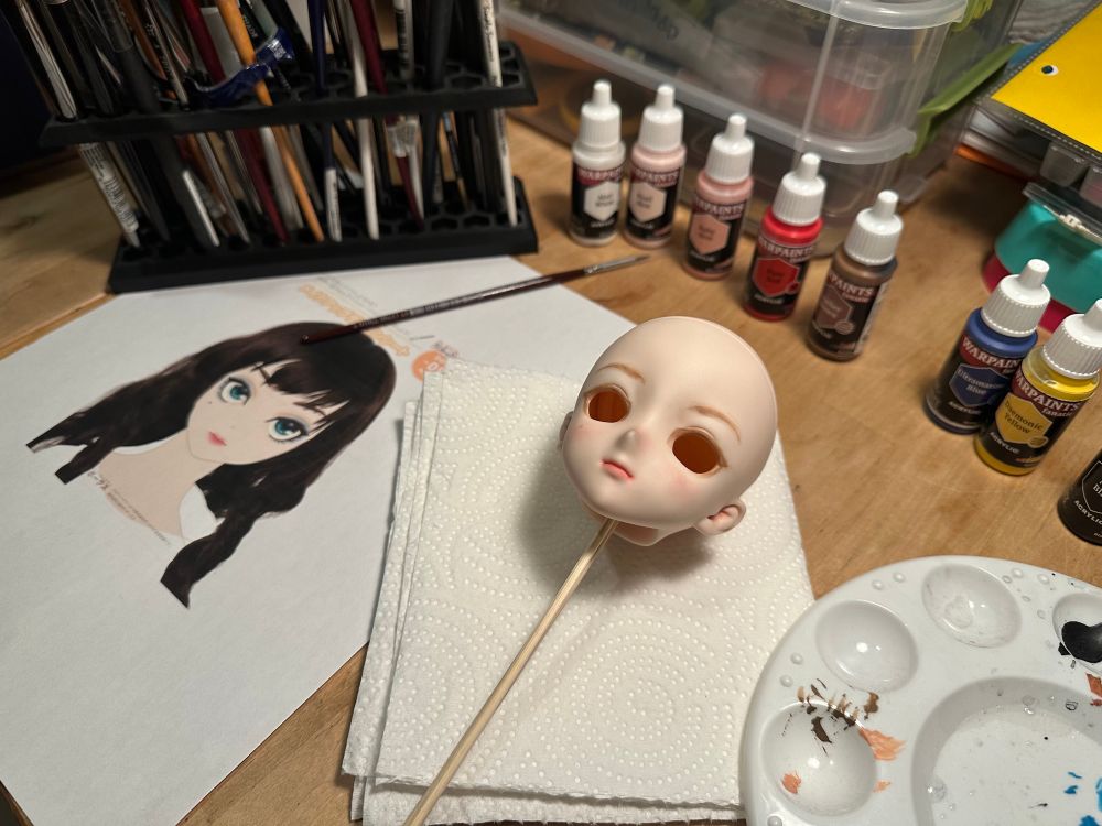 A half-painted doll head on a table, surrounded by paints and brushes 