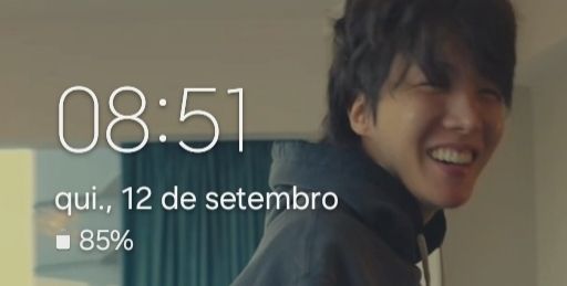Screen shot of a phone wallpaper. In it, Jhope of BTS (a young man), on the right of the screen, smiling. He's wearing a gray hoodie. Behind him, a blueish curtain, white ceiling and walls.