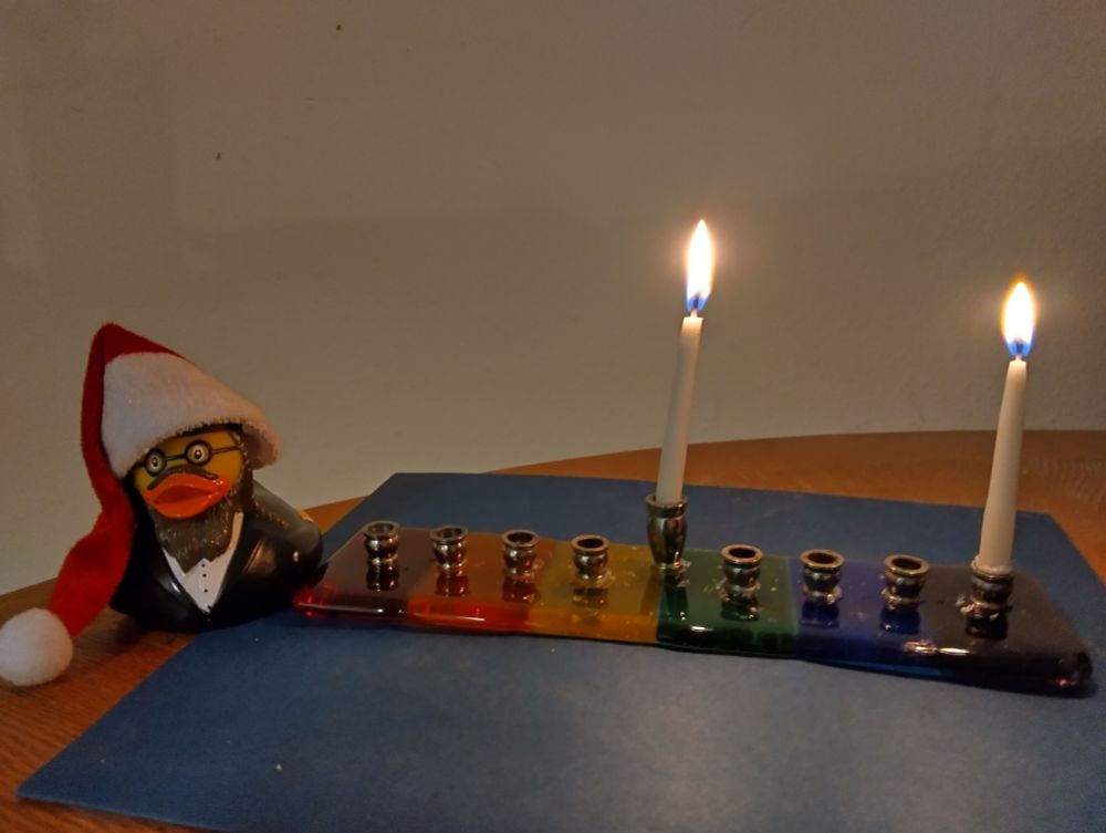A menorah in rainbow colors has the shamash lit and one candle for night one. A rubber duck that's dressed like an Orthodox Jew with payes and all also has a Santa hat on to recognize that other famous holiday 