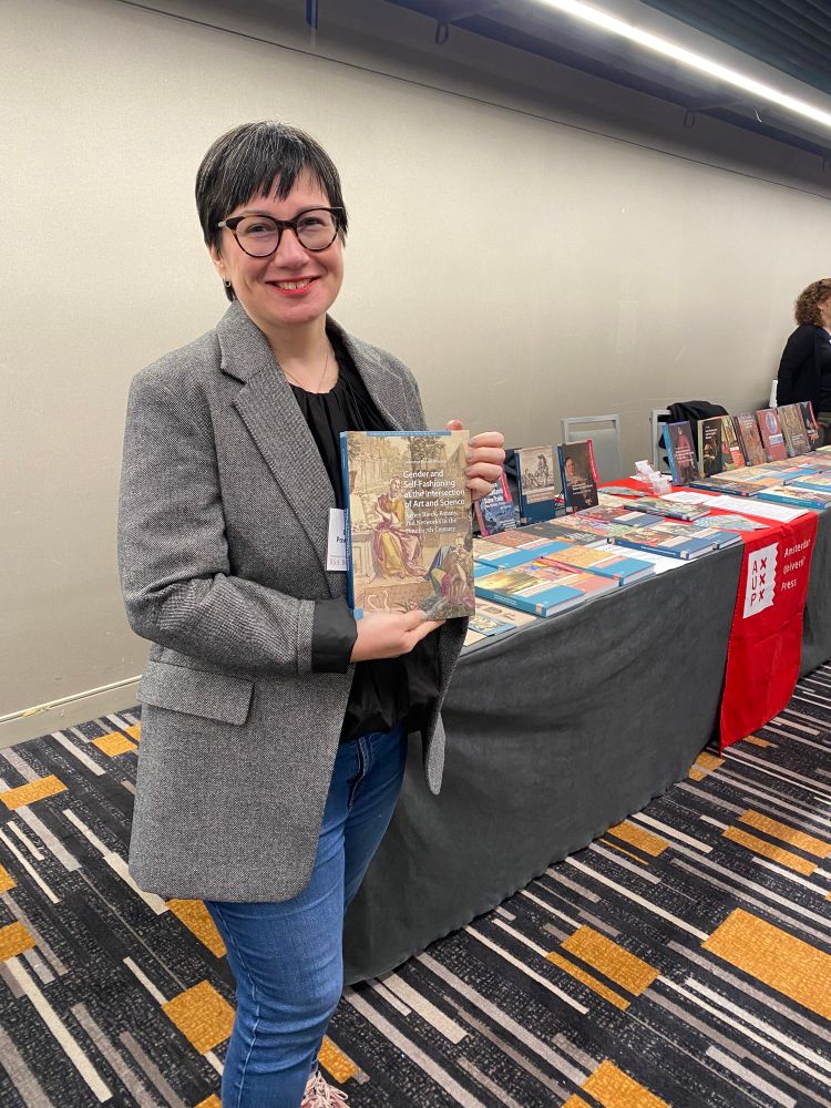 Catherine Powell-Warren holds up her book in front of table of books 