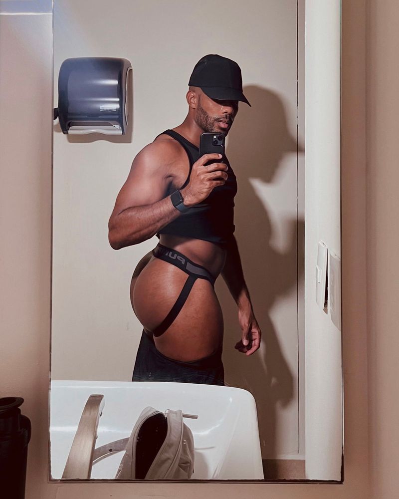 Muscle butt bottom taking a mirror selfie in a public bathroom. He's wearing a black cap, tank top and black shorts which are pulled down to expose a fat ass in a black jockstrap. The lighting casts a harsh shadow on the brim of the cap, obscuring his face. 
