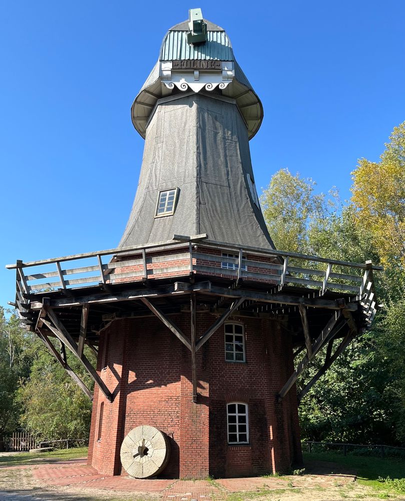 A windmill with two brick-built lower stories and a wooden superstructure but no sails. There’s a mill stone propped up against the wall. There’s autumn trees behind and a rich blue autumn sky. 