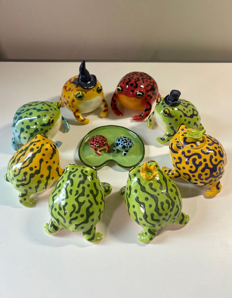 A group of 8 colorful ceramic frogs sit in a circle, surrounding and facing a ceramic lily pad dish with 2 tiny frogs sitting in it.