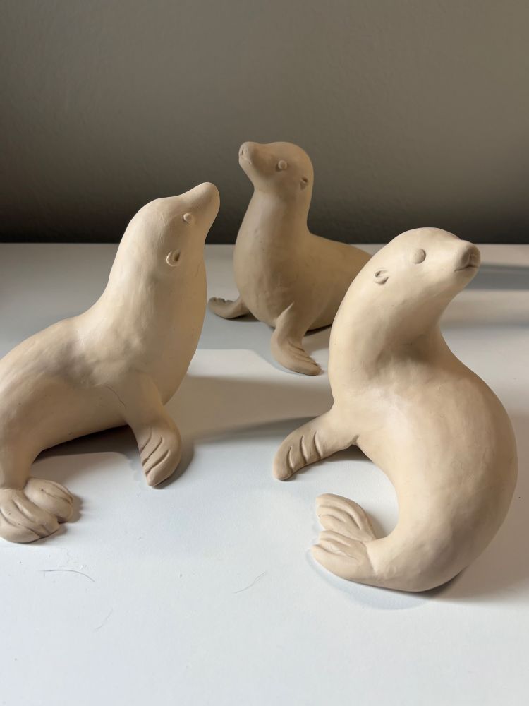 3 unfinished clay sea lion sculptures sit in a group, all slightly different poses.