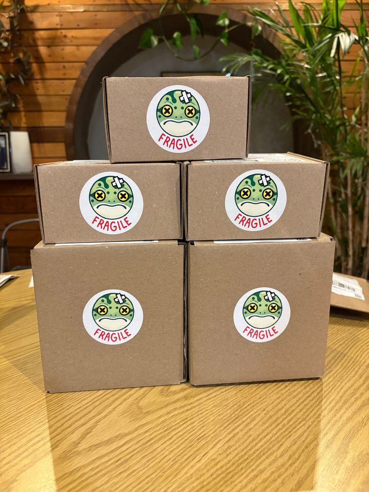 A pile of boxes with round stickers on them that read fragile. They have an image of a frog with a bandaid and x’s for eyes.