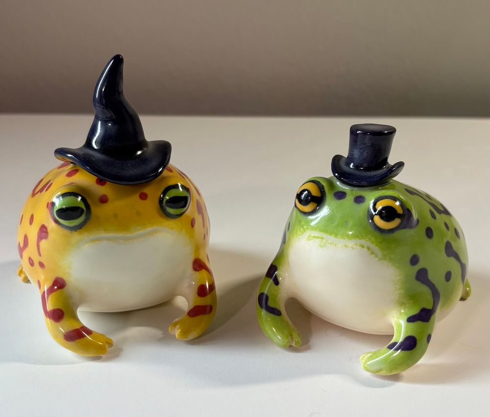2 ceramic frogs with hats. One is a witch hat, the other is a top hat.