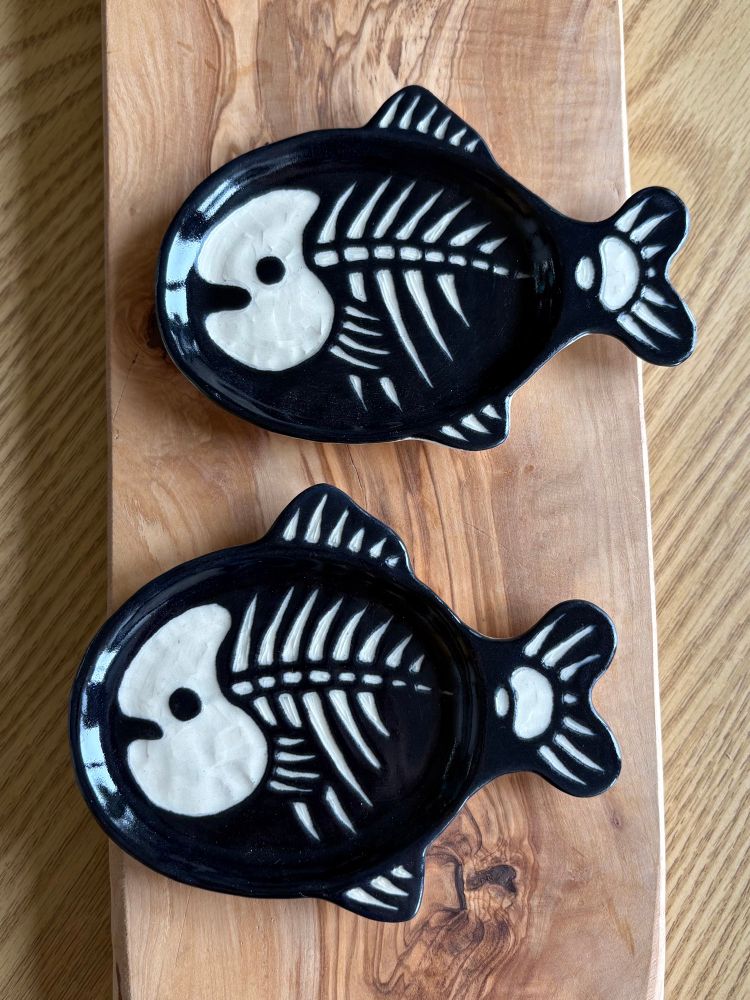 2 small fish dishes sit side by side. They are shaped like fish, painted black with a cute white skeleton design carved into it. They are coated with clear glaze.