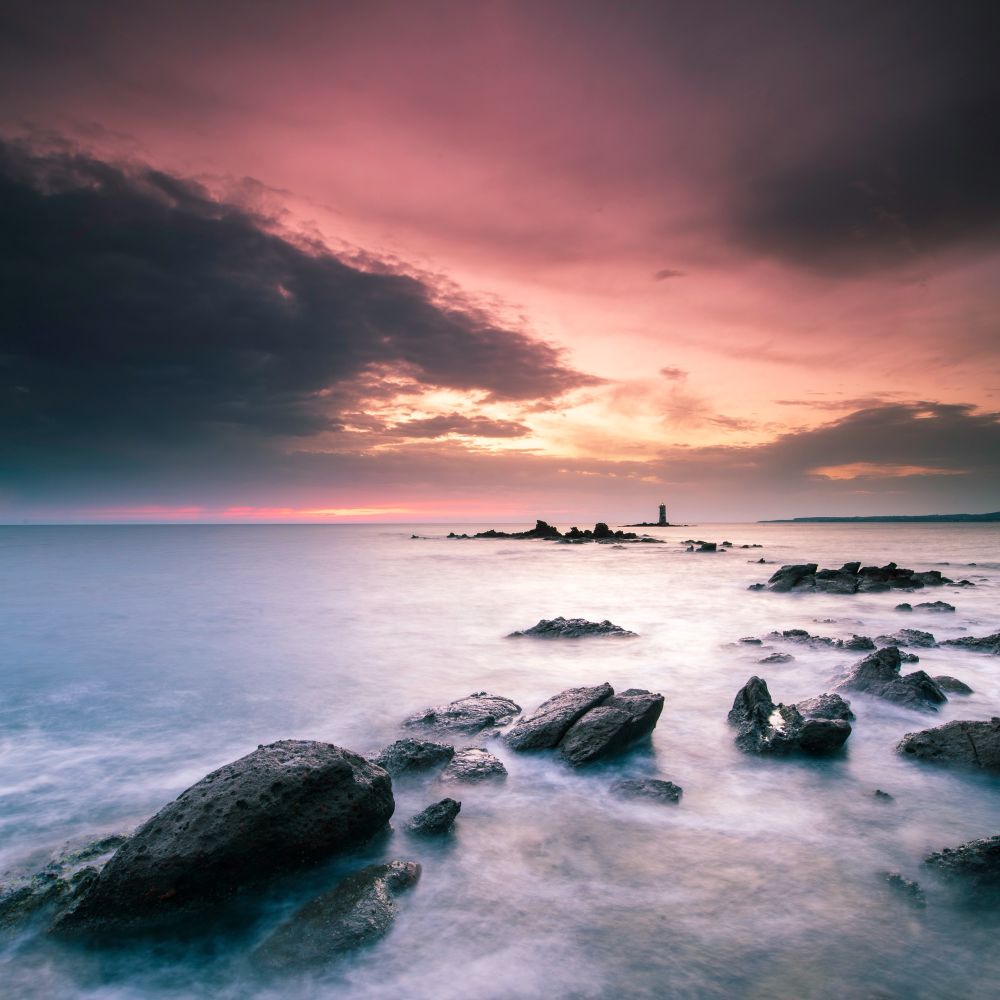 Rocks extending towards the horizon with a tiny lighthouse barely visible in the distance during a sunset witnessed at the shore of a Sardinia beach. 