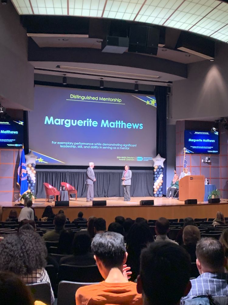 Photo of an auditorium stage showing the name Marguerite Matthews for Distinguished Mentorship on a big screen at the NINDS Director’s Award Ceremony 