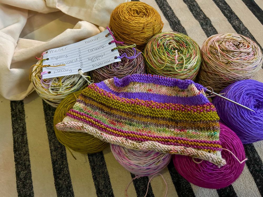 A striped scarf sits on top of caked yarns that were used to make the stripes. An index card with yarn scraps next to the genres the yarns represent sit beside the scarf in progress. An empty canvas tote bag is beside the yarn. It all sits atop a black and cream striped blanket. 
