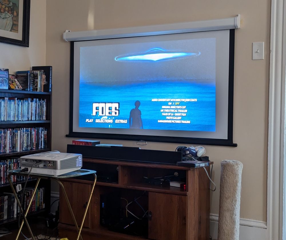 A living room projector screen showing the DVD menu for the movie FOES, with a silver flying saucer over the ocean and a silhouetted man watching it.