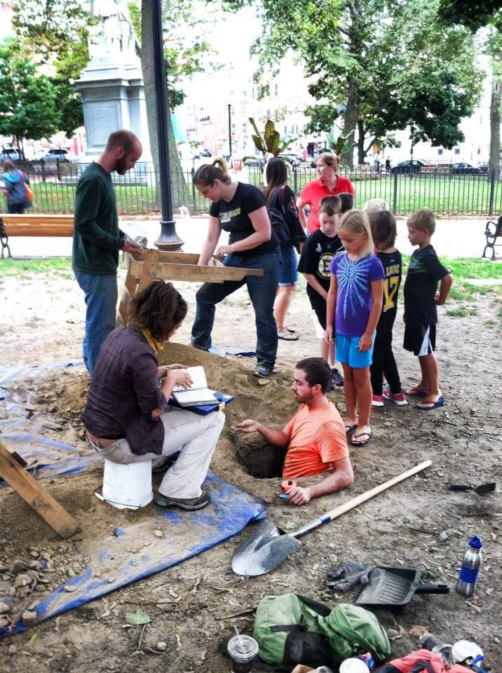 A group of 4 volunteers digging an archaeological shovel test pit and screening the contents beside the pit as 6 children look on.