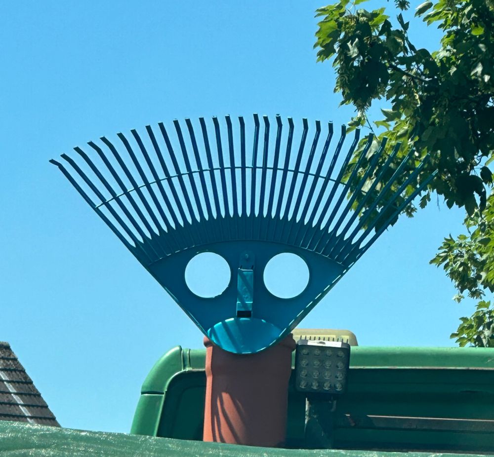 Türkiser Rechen mit zwei kreisrunden Aussparungen, der in einem Rohr auf der Ladefläche eines Lasters steckt. Rechts ragen Äste ins Bild. 