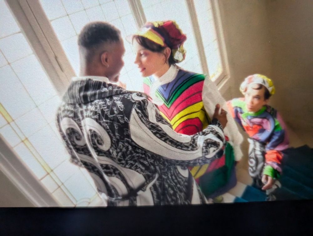 3 people in super colorful graphic outfits and hats on interior stairs as part of the Paris Olympics opening ceremonies
