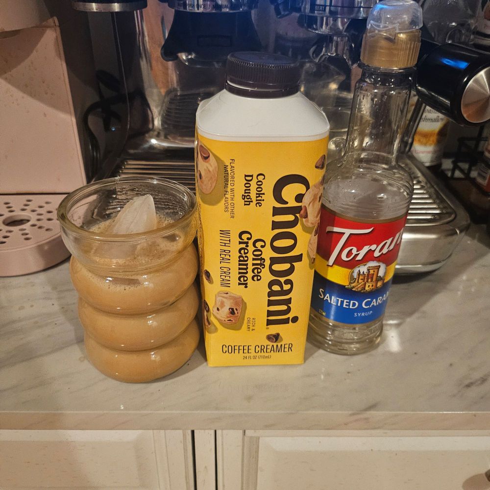 An iced latte sitting next to chobani cookie dough creamer and salted caramel syrup