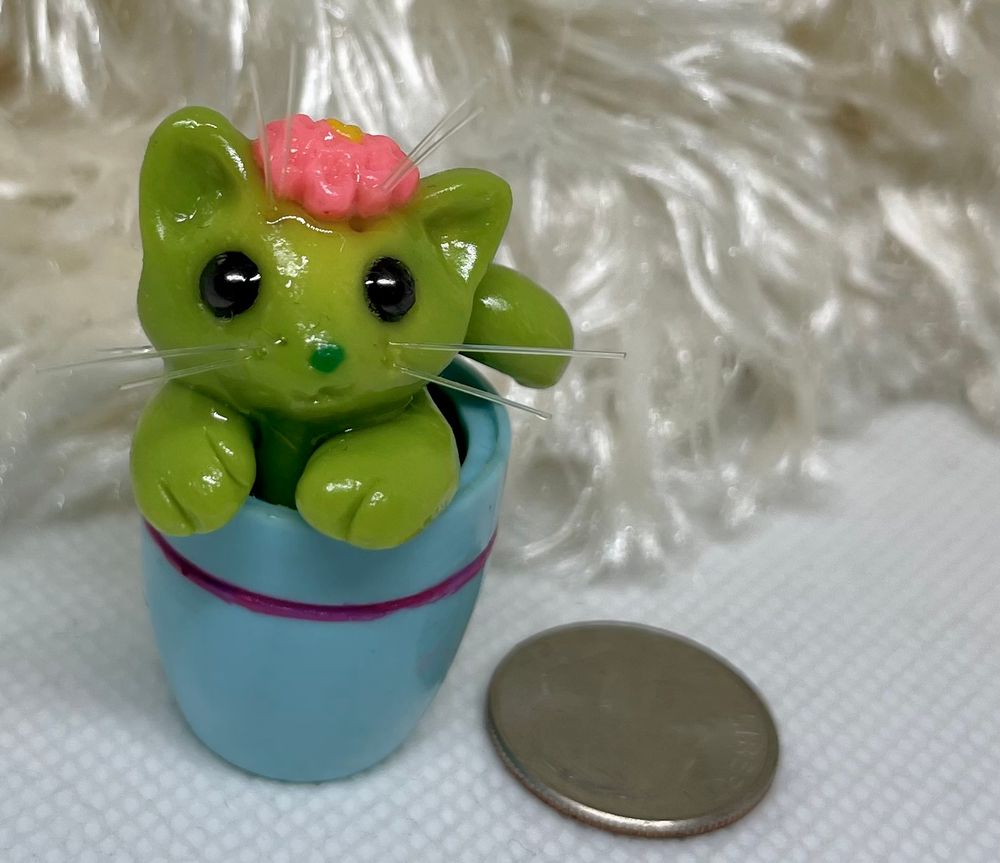 A polymer and resin miniature sculpt of a green cat with flexible plastic/line bristles. The cat resembles a cactus with a pink cactus flower atop their head, and is perched inside an aqua planter. US Quarter coin shown for size reference