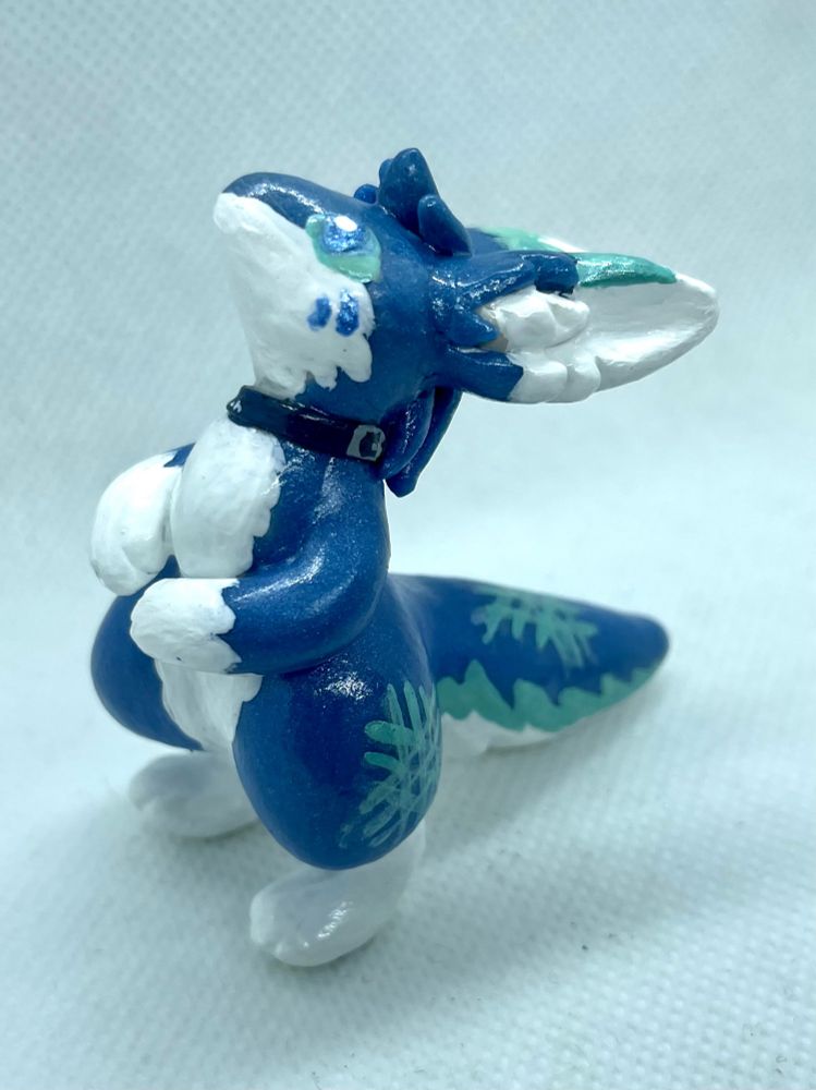Polymer clay miniature figurine of Largen, a blue-and-white foxlike critter with glow markings on his flank and tail. He is standing in his hind legs with his hands folded in front of him.

Seen from left. US Quarter coin for size reference.